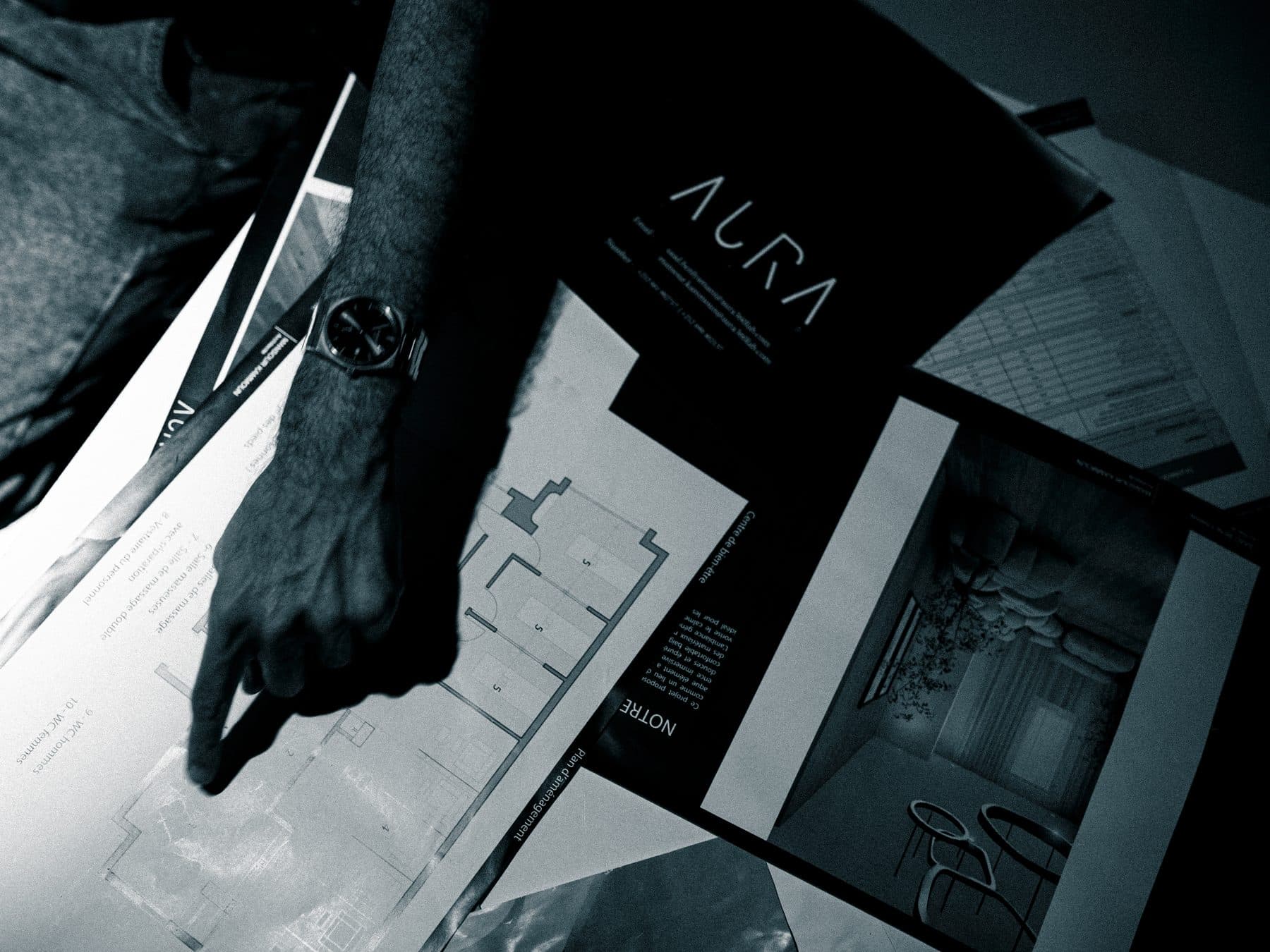 Hands pointing at AURA360LAB floor plans and renders on a desk